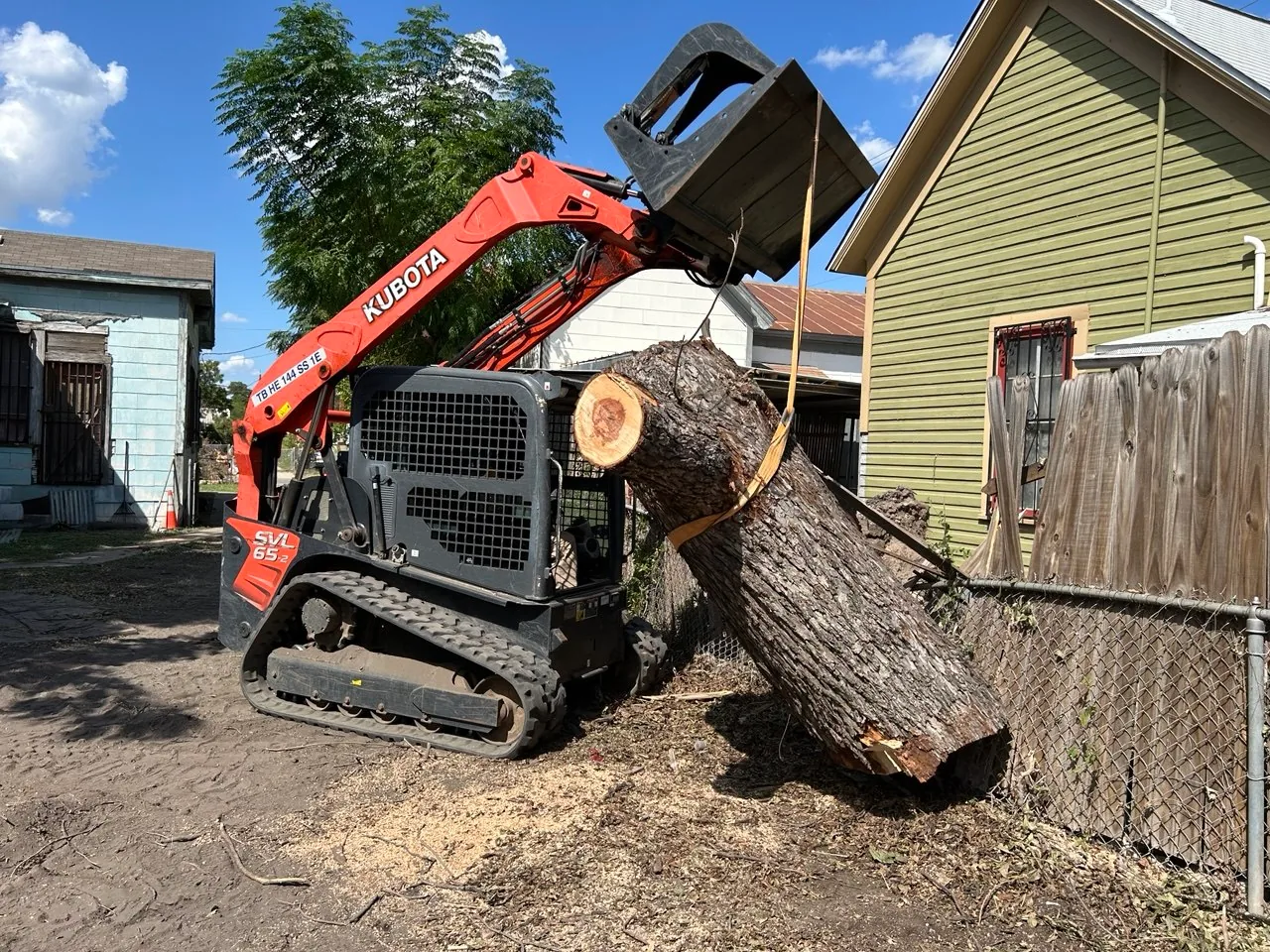 Skid steer lifting a large cut tree trunk between homes during San Antonio storm cleanup.